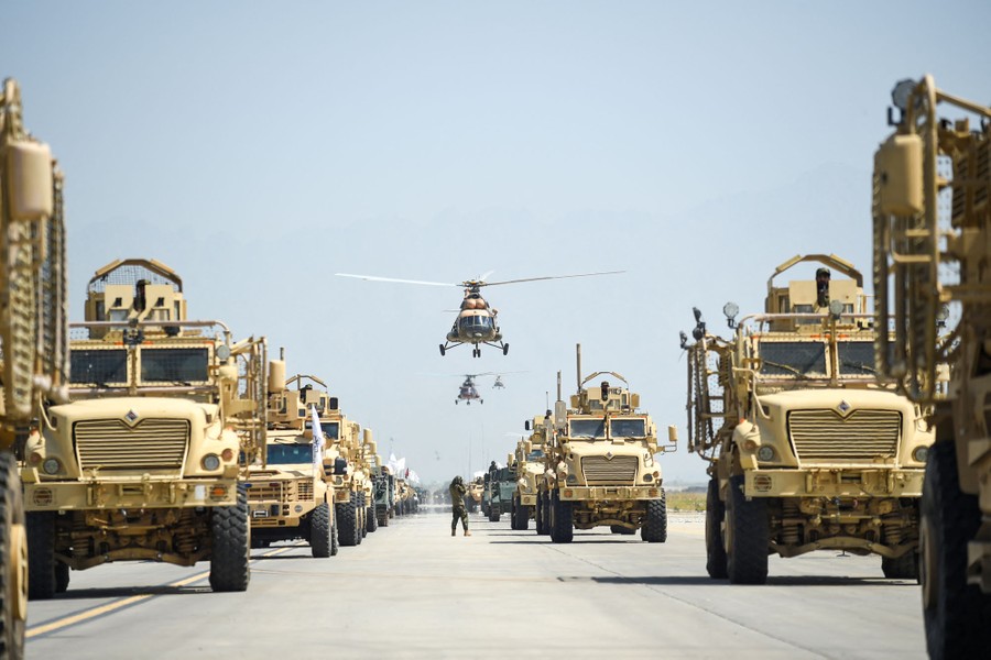 Dozens of military vehicles drive in a parade beneath several helicopters.