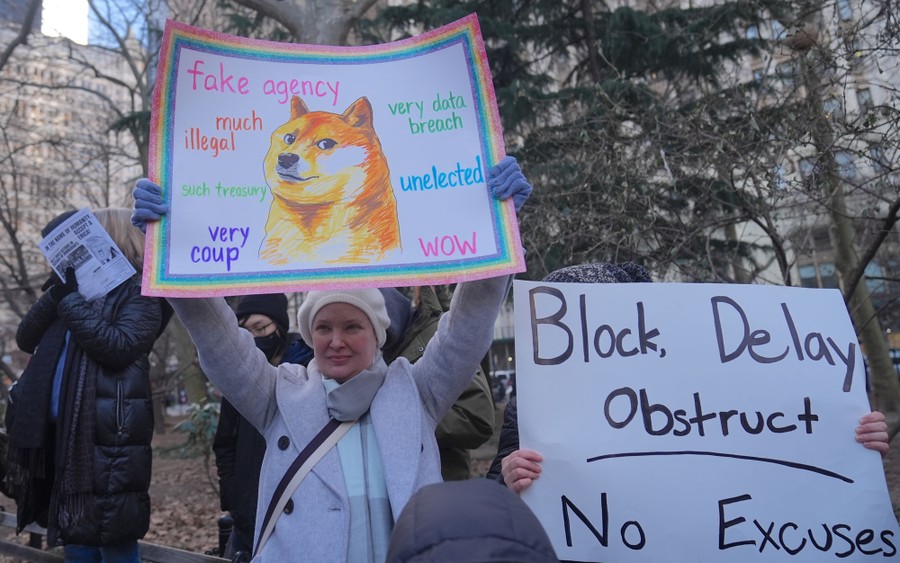 Several people, part of a protest group, hold signs. One sign depicts a famous Shiba Inu dog surrounded by words reading "much illegal," "fake agency," and "very data breach." The other sign reads "Block, delay, obstruct. No excuses."
