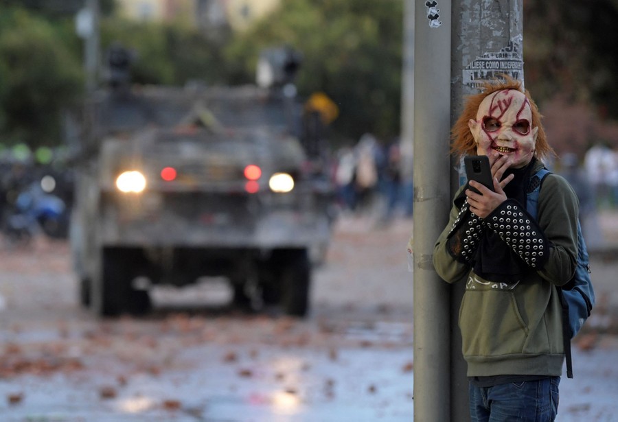 A protester wearing a scary rubber mask stands behind a pole on a street as riot police are seen in a vehicle in the background.