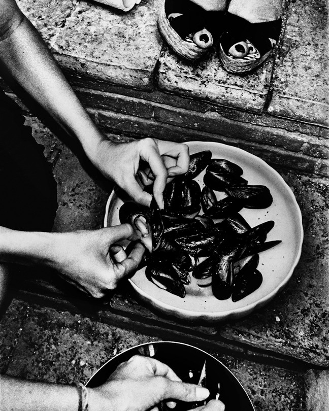 A black-and-white photo of someone cleaning mussels, with someone else's shoes in the photo, in black strappy sandals
