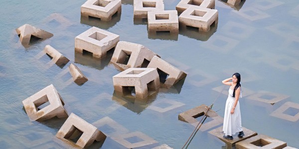 A woman stands on a cube-shaped concrete block, among many such blocks piled haphazardly in shallow water.