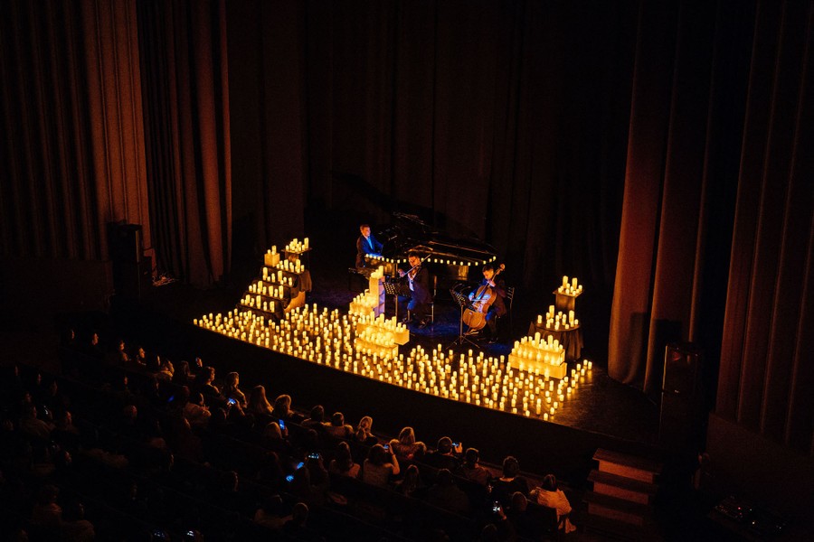 A trio of musicians plays on a stage filled with candles, before an audience.