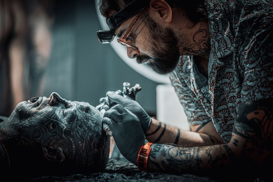 A tattoo artist leans over, applying ink to the head of another person.