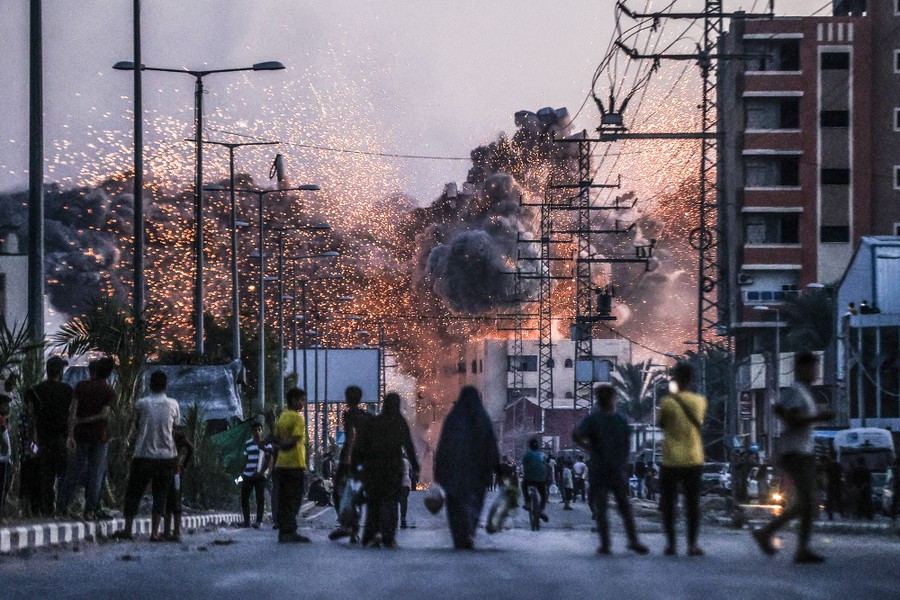 People in a street look on as a building in the distance explodes, struck by a missile.