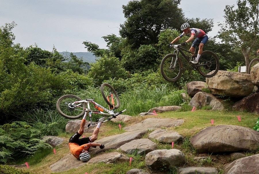 A mountain-bike rider crashes among large rocks as another racer approaches from above.