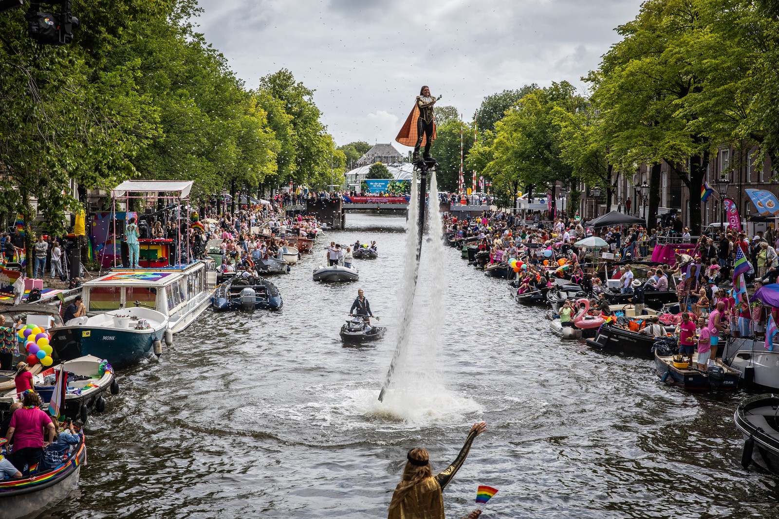 Festival-goers in many boats watch a person performing on a flyboard in a canal.