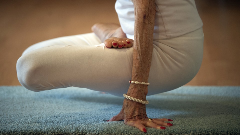 An elderly white person in white clothing with red nail polish does an exercise on a blue mat.