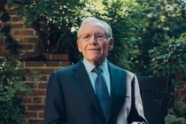 An older man in a blue suit and tie standing in front of a brick wall and lush greenery