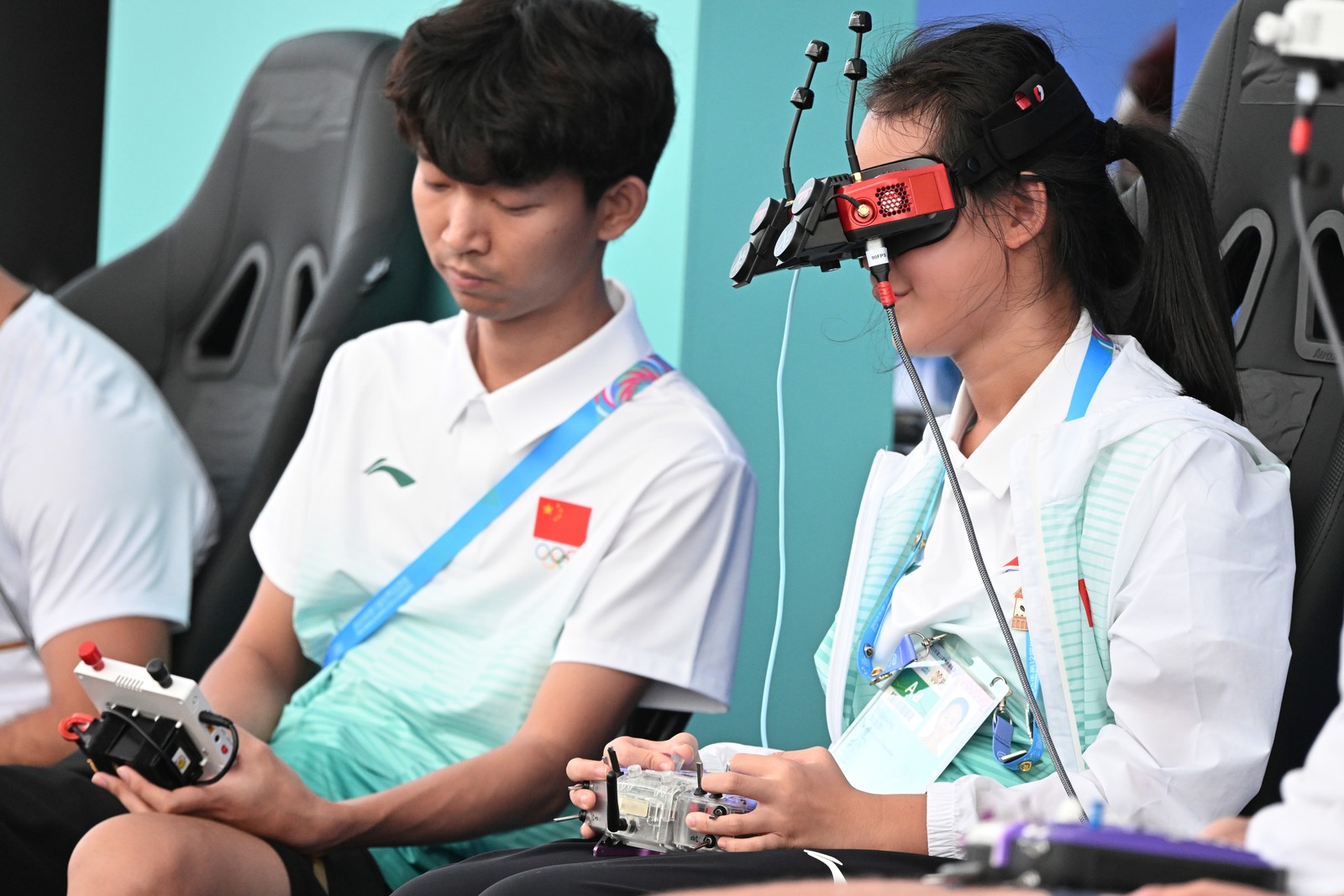 Two people sit side-by-side, one wearing a complicated looking headset and using a remote control, during a drone race.