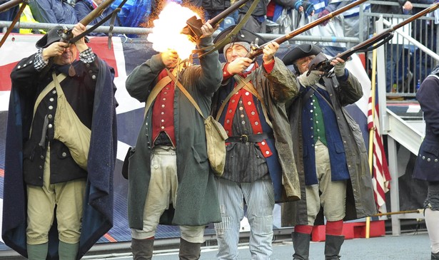 Six men dressed in reenactment costumes firing muskets. An audience in football jerseys and rain ponchos sit behind them.