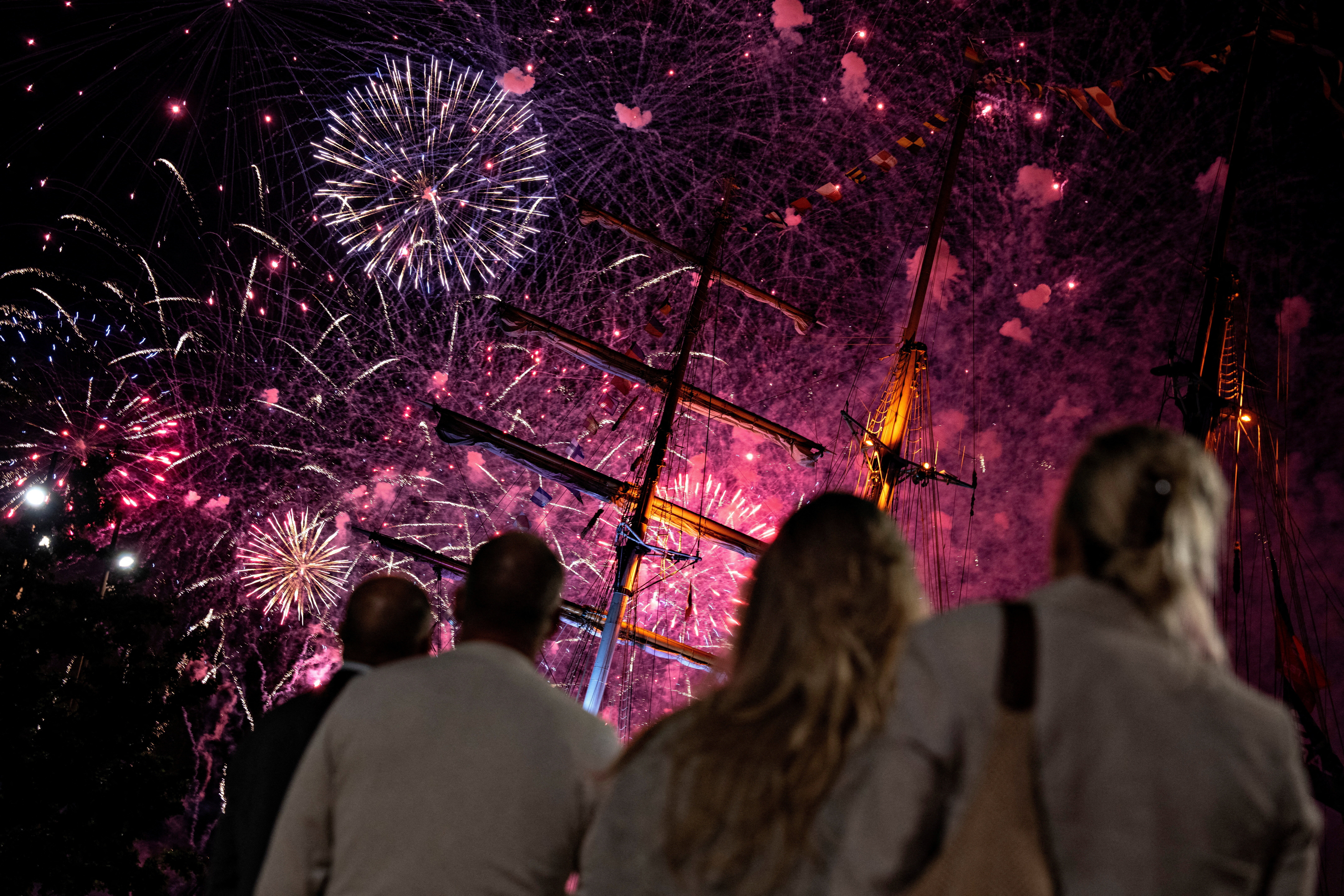 People watch fireworks explode above tall ships.
