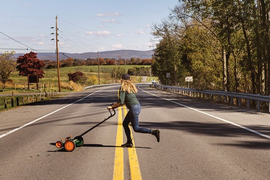 photo of woman with long hair pushing hand mower across rural two-lane road with mountains in background