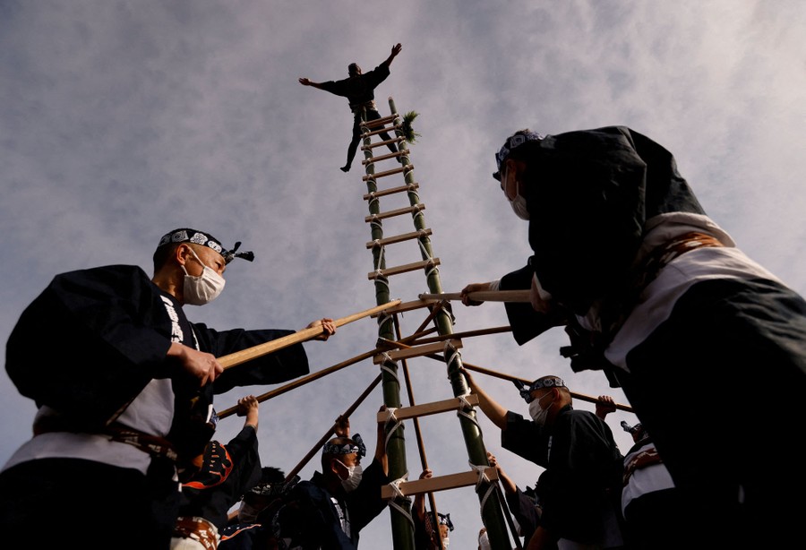 Several men in traditional clothing hold a bamboo ladder upright, with another man on top, balancing on his stomach.