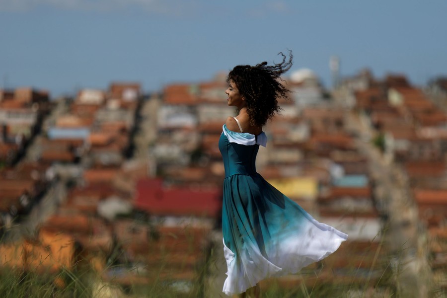 A young woman wearing a flowing dress stands on a hillside in front of buildings on a sunny day.