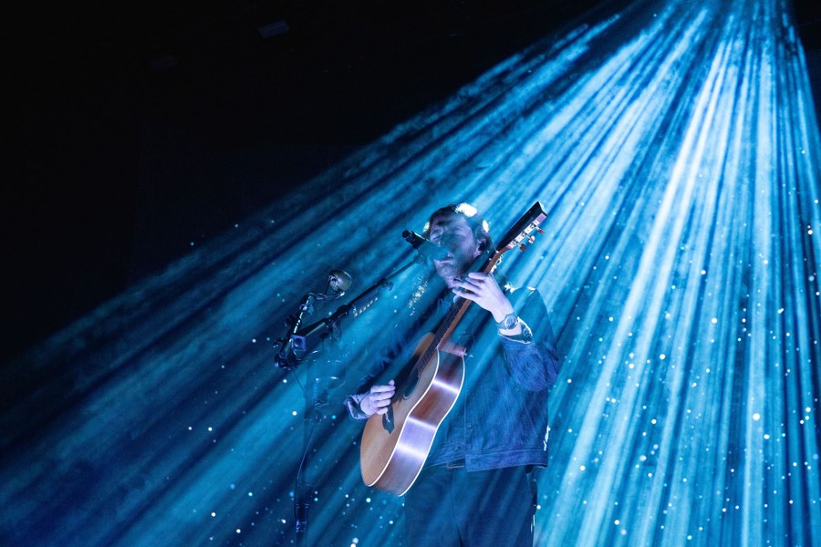 A performer plays guitar onstage, beneath streams of light.