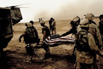 Soldiers on a barren field lift a body on a stretcher, covered in an American flag, into a helicopter.