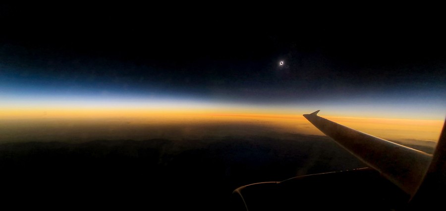 The eclipsed sun appears like a small circle in a darkened sky above a strip of clouds and the horizon, seen from an aircraft window.