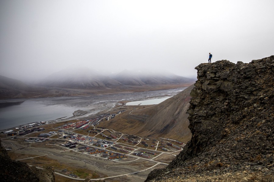 A person stands on a cliff above a small town, seen below in the distance.