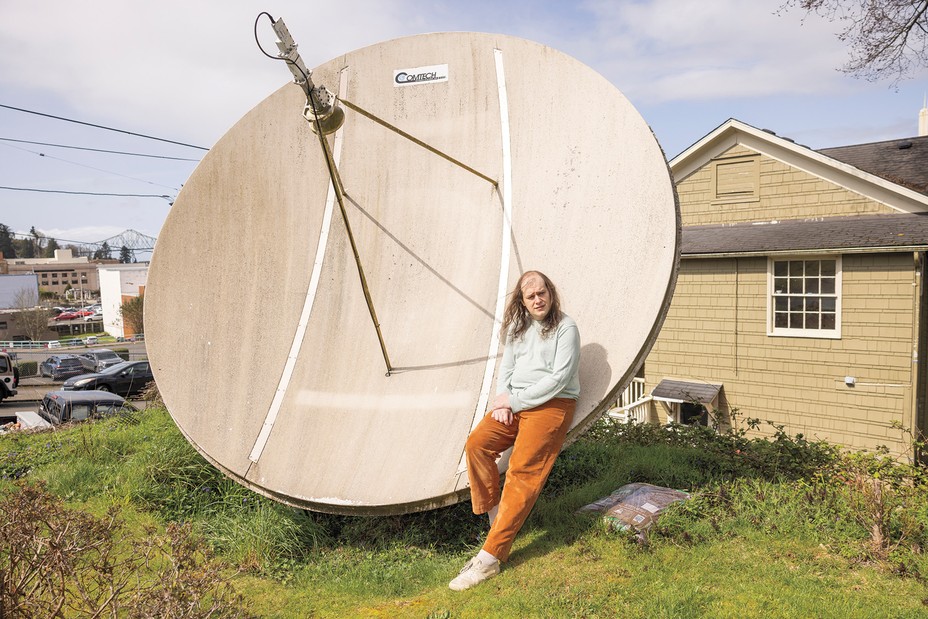 photo of long-haired person leaning against large satellite dish with houses in background