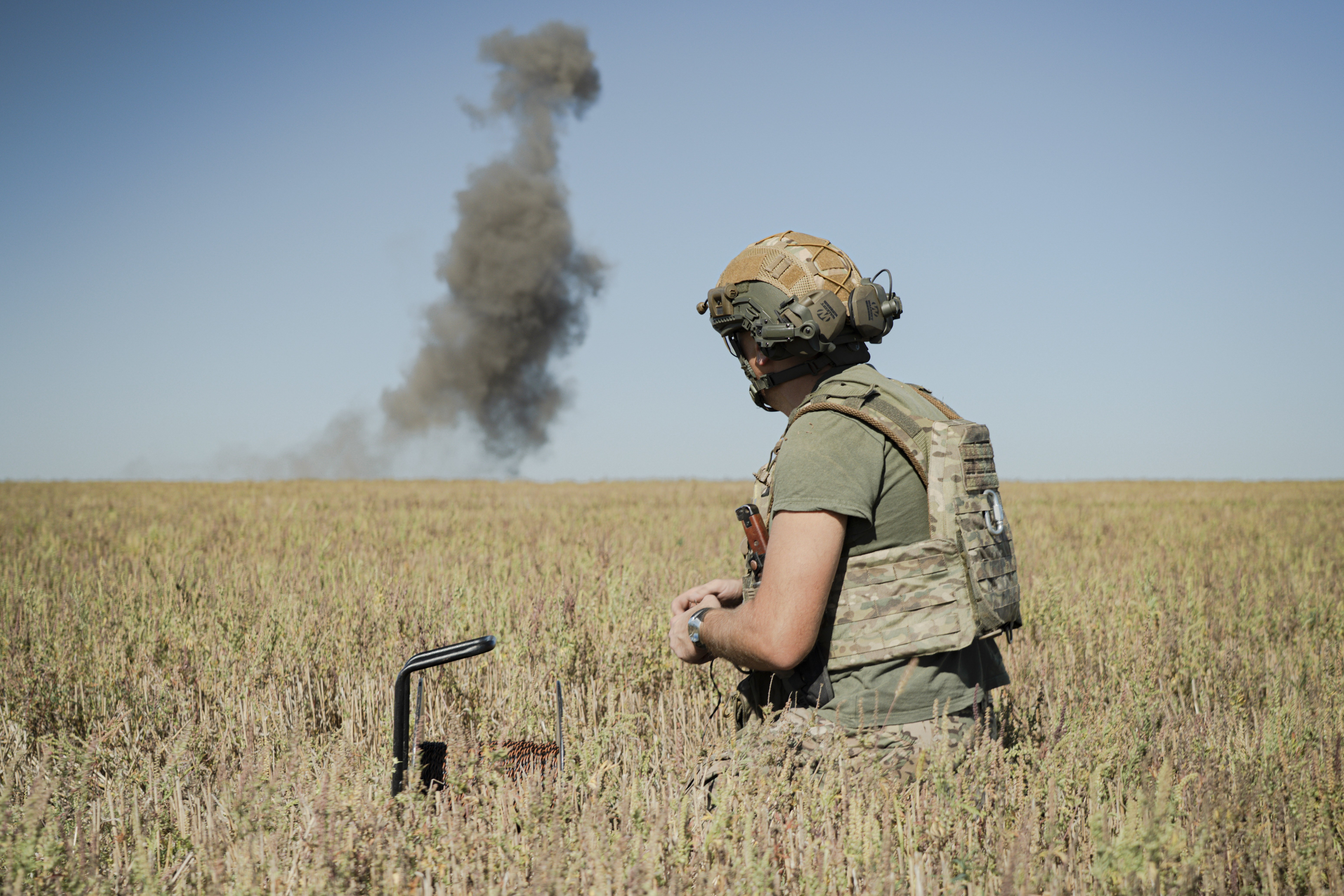 A soldier kneels in a farm field, as smoke rises from a distant explosion.