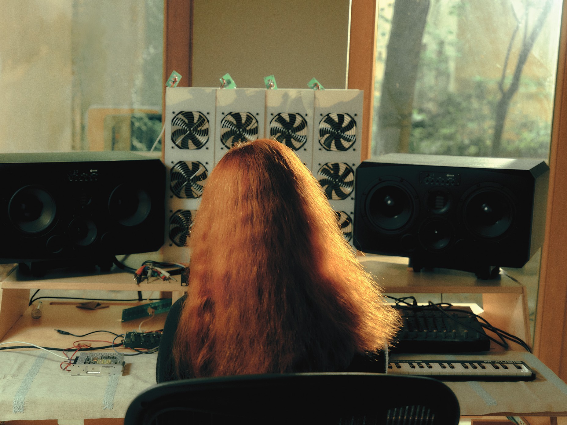 photo from behind of woman with long red hair sitting at desk with two speakers, midi keyboard, and electronic spare parts with window in background