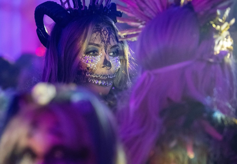 A person wearing horns and crystals on their face is framed by other costumed revelers.