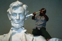 A person uses a cloth-wrapped pole to clean the ear of a large statue of Abraham Lincoln.