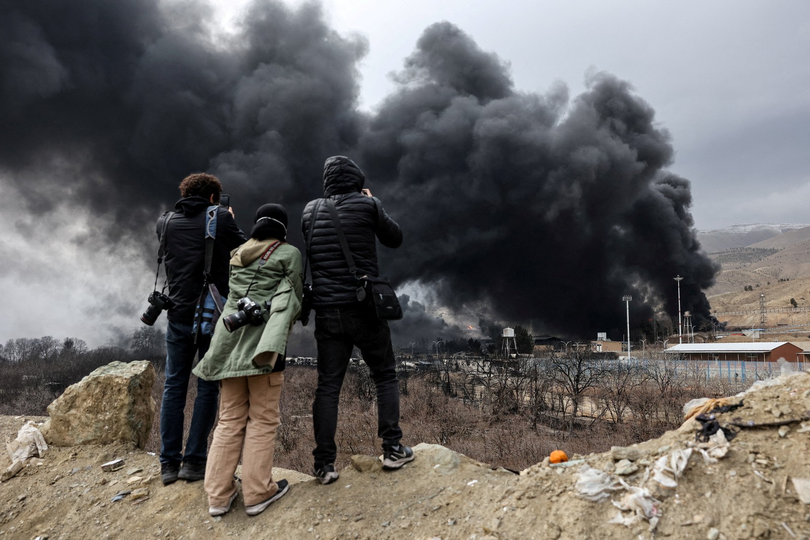 Three people stand on a hill photographing rising black smoke from a nearby fire.