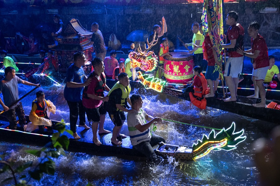 Several dragon boats pass each other in a narrow canal, with adults and children playfully riding and splashing.