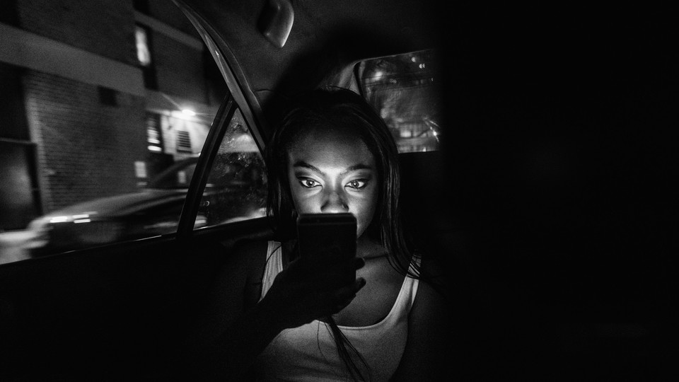 A passenger looks at their phone in the backseat of a dark car.