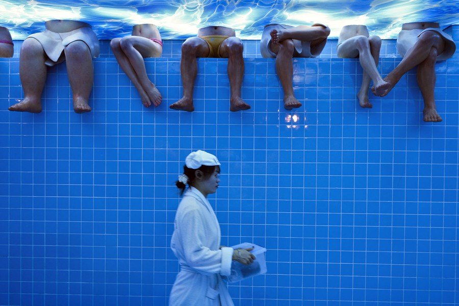 A waitress walks past a blue-tiled wall topped with the legs of several mannequins; she seems to be walking along the bottom of a swimming pool.