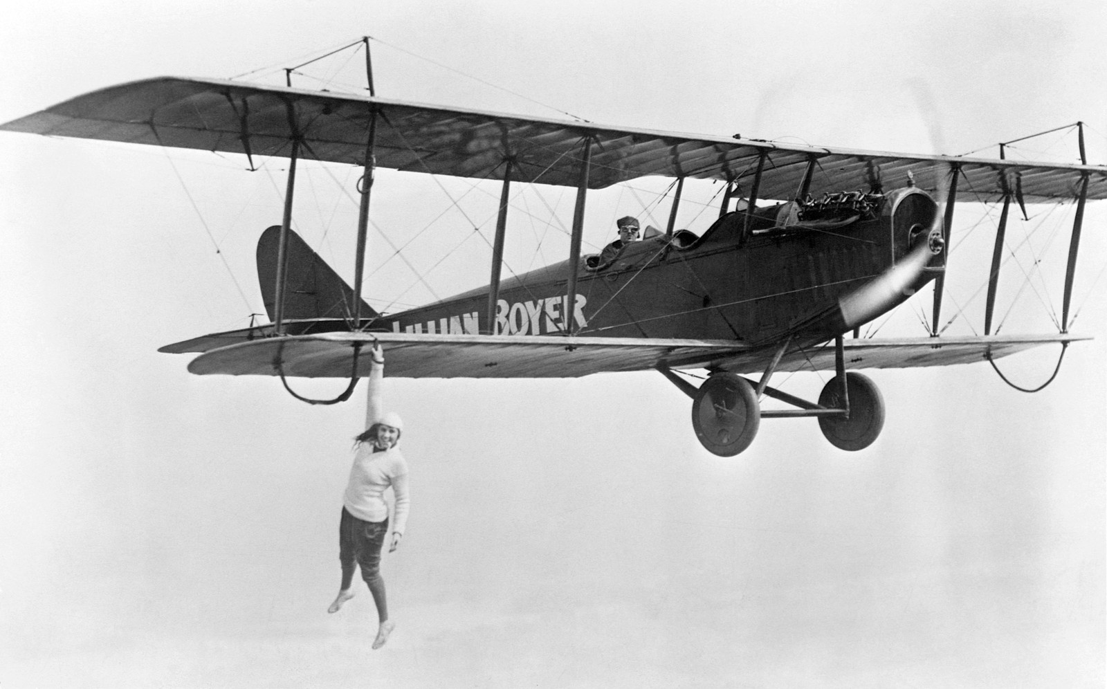 A stunt performer hangs from the bottom wing of a biplane, using only one hand.