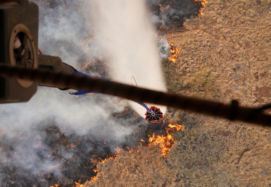 A view, looking down from inside a helicopter, of a collapsible water bucket dangling from a cable, as it releases water onto a wildfire below