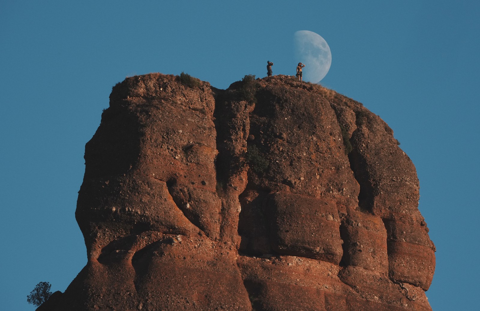 Two climbers stand atop, a monolithic rock formation, with the moon visible in the blue sky behind them.