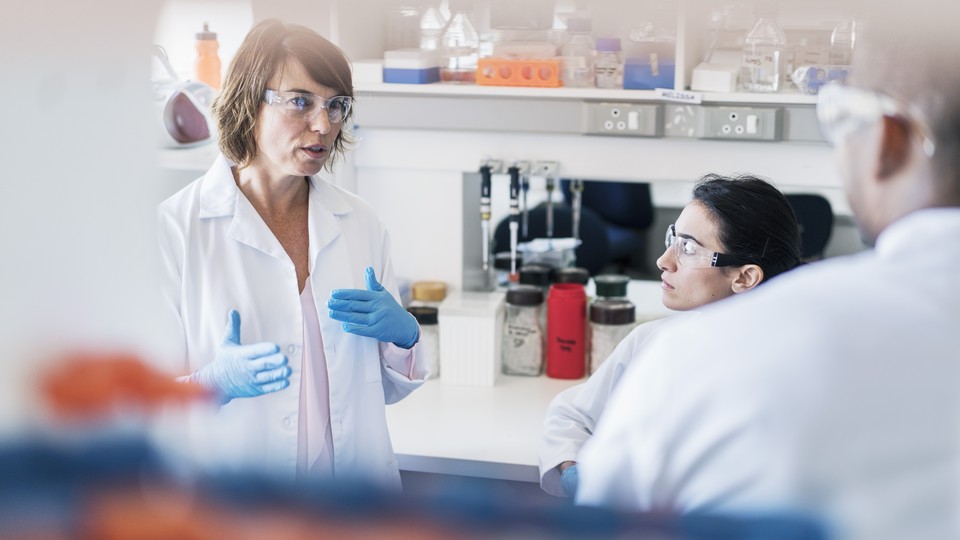 A woman wearing a lab coat, lab goggles, and plastic gloves, speaks and gestures.