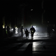 Color photo of a Cuban street in the dark, showing a the silhouettes of people lit up by headlights.