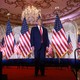 Donald Trump stands on a red carpeted stage, flanked by American flags.
