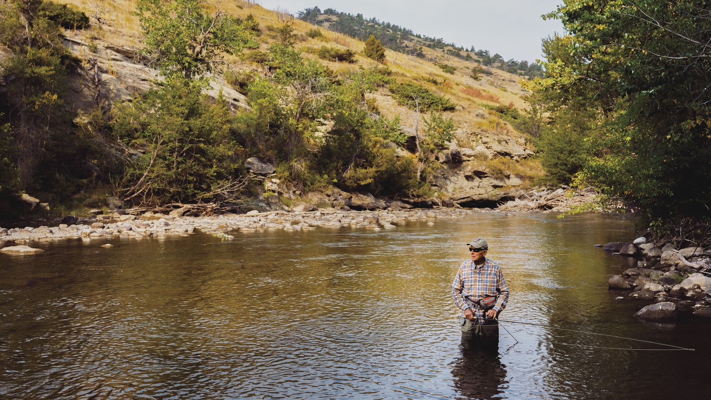 Color photo of a man in waders and a plaid shirt standing knee-deep in a stream holding a fly-fishing rod with hills in the background.