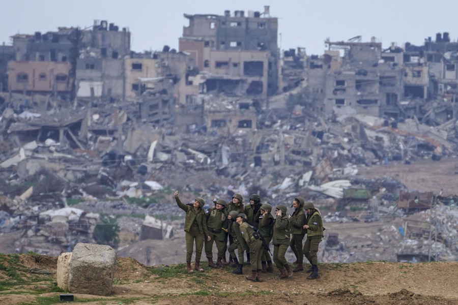 Israeli soldiers pose for a selfie on the Gaza Strip border, with destroyed buildings in Gaza in the background.