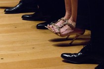 A photo depicting a row of men's shoes and one woman wearing heels