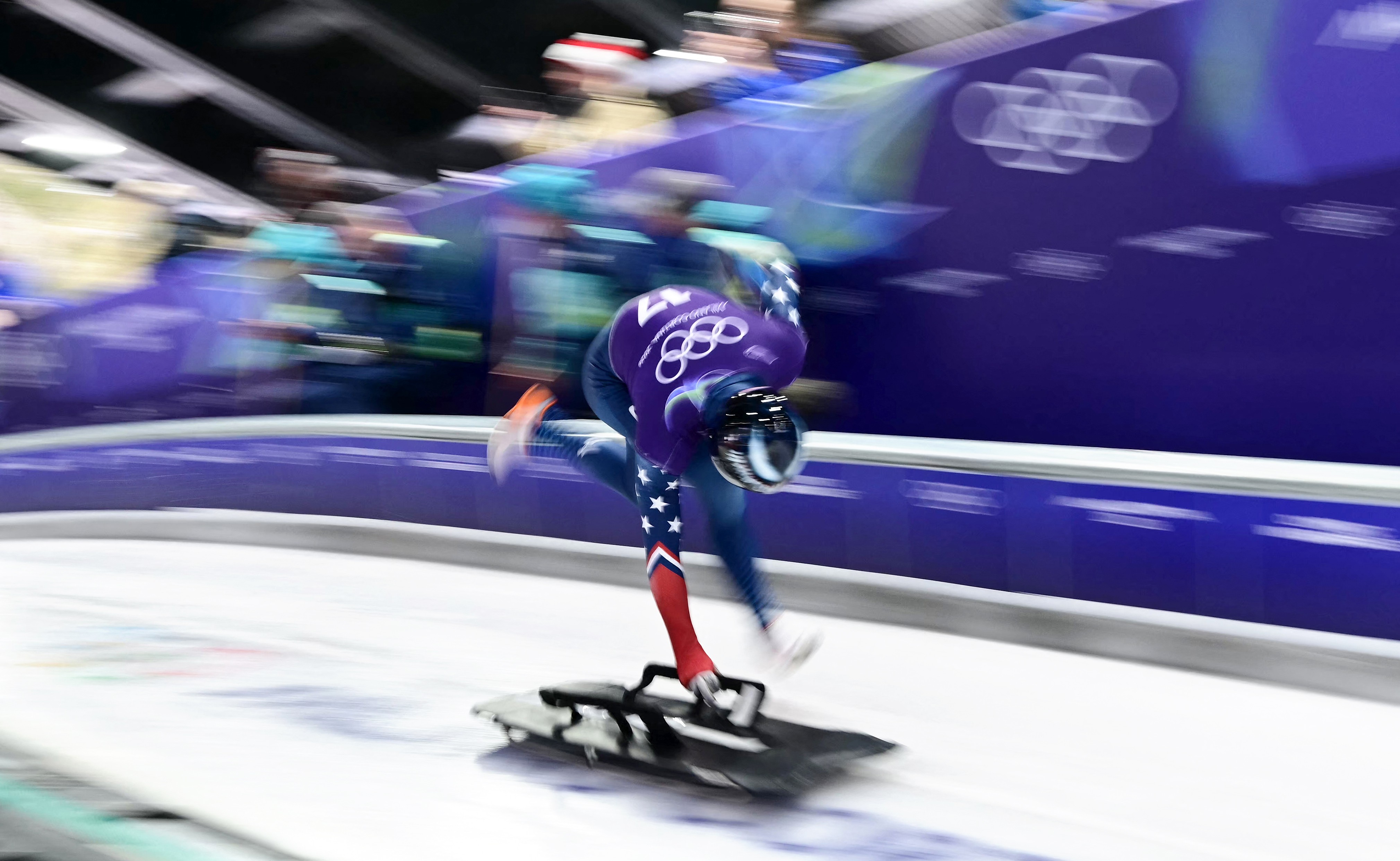 An Olympic athlete sprints on an ice track, bent over, pushing a small sled at the start of a run, legs and arms blurred by motion.