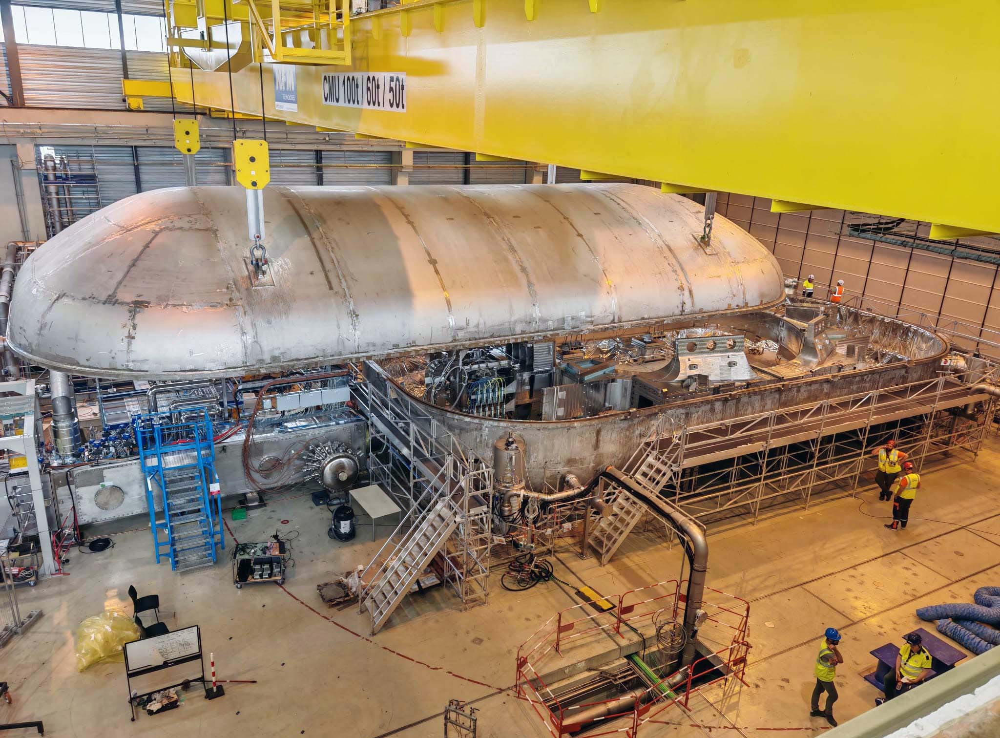 A huge, rounded metal lid is suspended beneath a gantry crane, about to be placed on top of a chamber filled with complicated equipment.