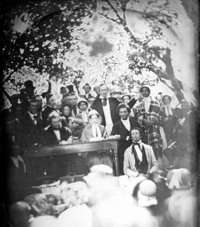A grainy black and white photo of the convention with people standing around, including Frederick Douglass, Gerritt Smith, Mary Edmonson, George W. Clarke, Theodosia Gilbert, Emily Edmonson, and James Caleb Jackson
