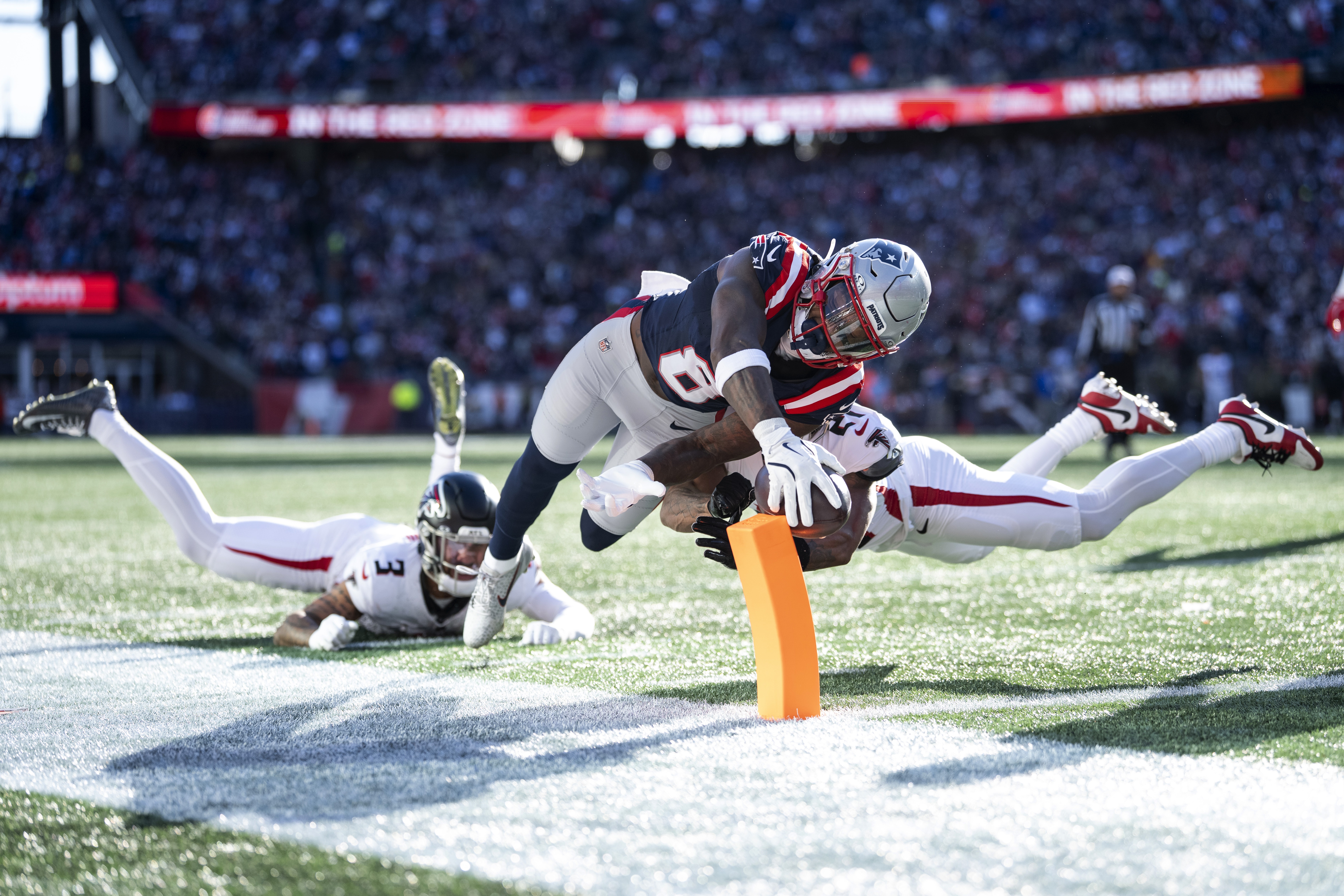 A football player dives for the end zone, holding the ball out and hitting a small soft pylon on the boundary line.
