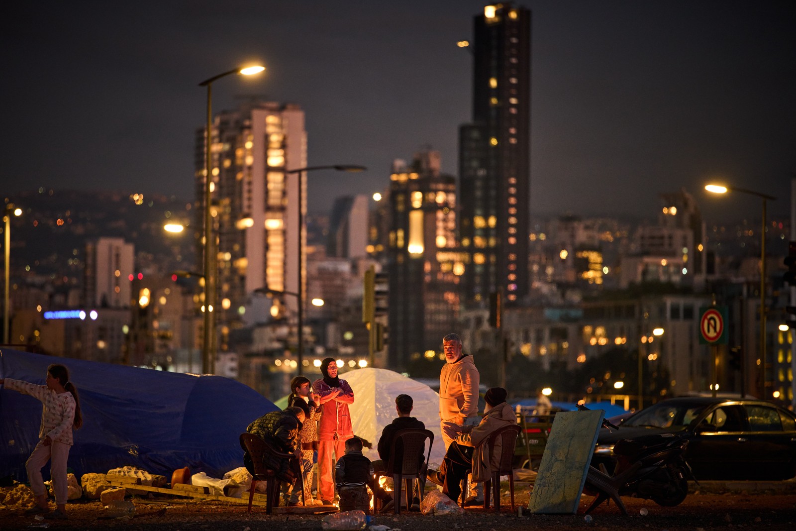 A family gathers around a fire, outside of tents, with city buildings visible in the background.