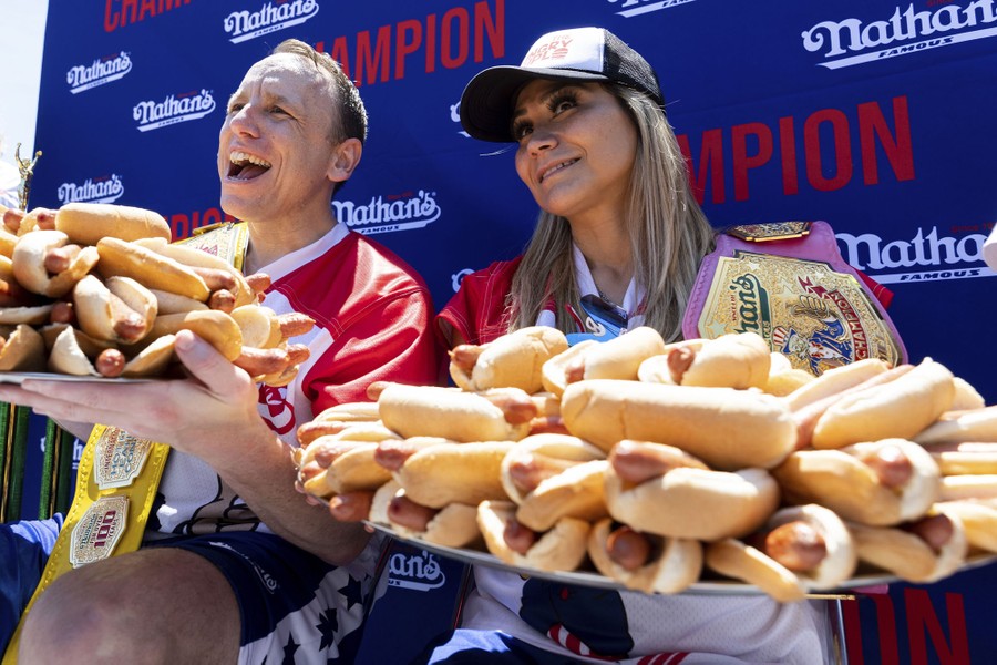 A man and a woman pose while holding up platters overflowing with dozens of hot dogs.