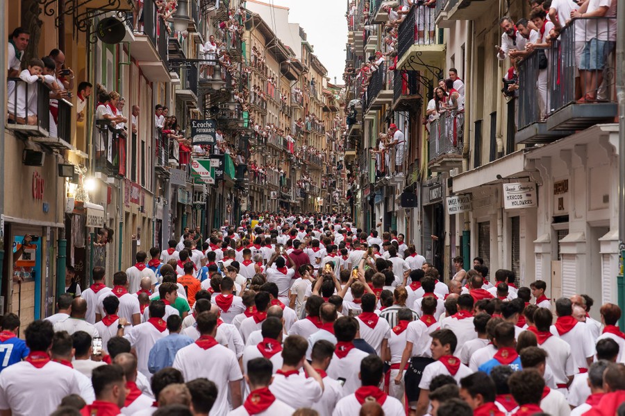 A crowd of people, most wearing white T-shirts with red bandannas around their necks, fills a street surrounded by tall buildings, waiting. for a bull run to begin.