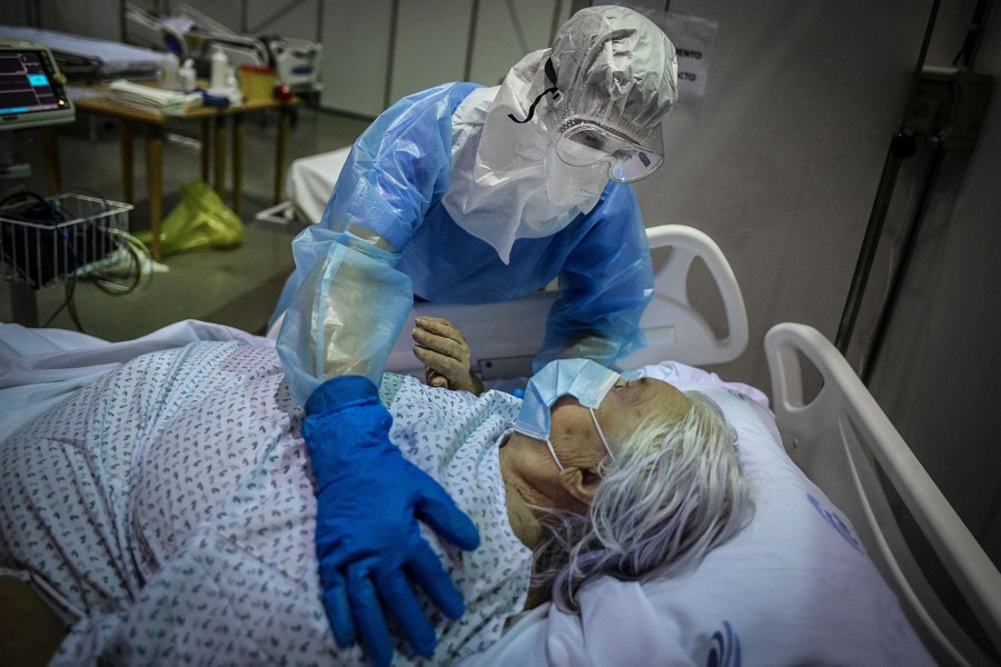 A health-care worker wearing protective equipment leans over a patient, holding their shoulder as they lie in a hospital bed.