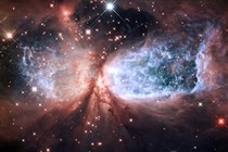 Gas, dust, and starlight form a glowing explosion, vaguely shaped like an hourglass lying on its side.