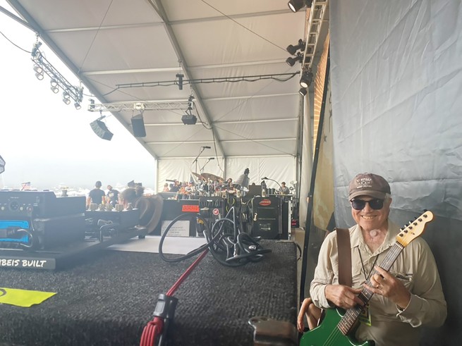 photo of smiling man wearing sunglasses and baseball cap standing and holding a green guitar on the side of a huge festival stage, with large band and musical equipment in background